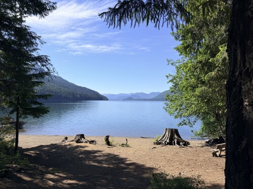 A sandy beach bordered by tall evergreen trees frames a clear alpine lake with distant mountains and blue sky.