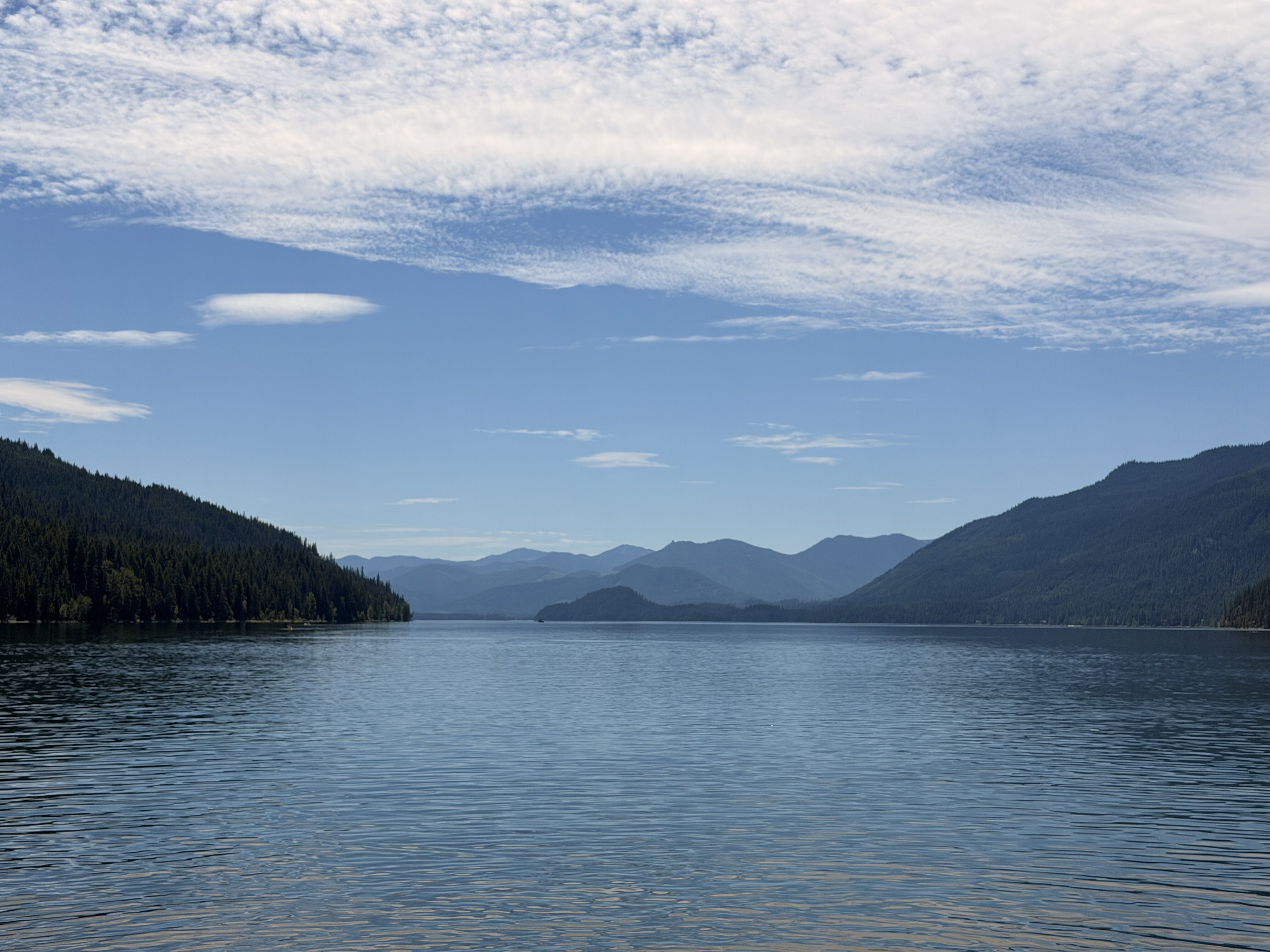 A calm mountain lake surrounded by dense evergreen forests and distant snow-capped peaks under a partly cloudy blue sky.