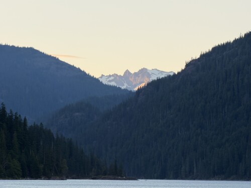A snow-capped mountain peak rises above a densely forested valley with a still lake in the foreground, captured in muted blue and beige tones.