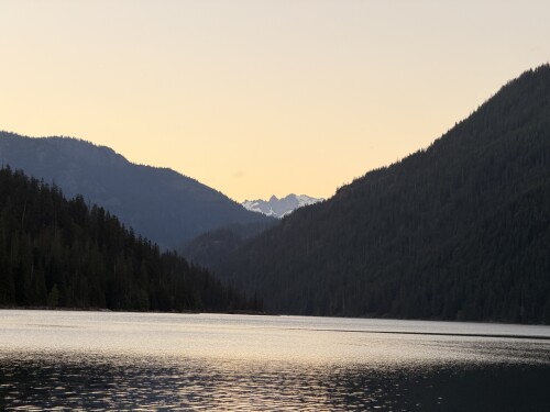 A calm mountain lake surrounded by forested peaks at dusk, with snow-capped mountains visible in the distance under a pale sky.