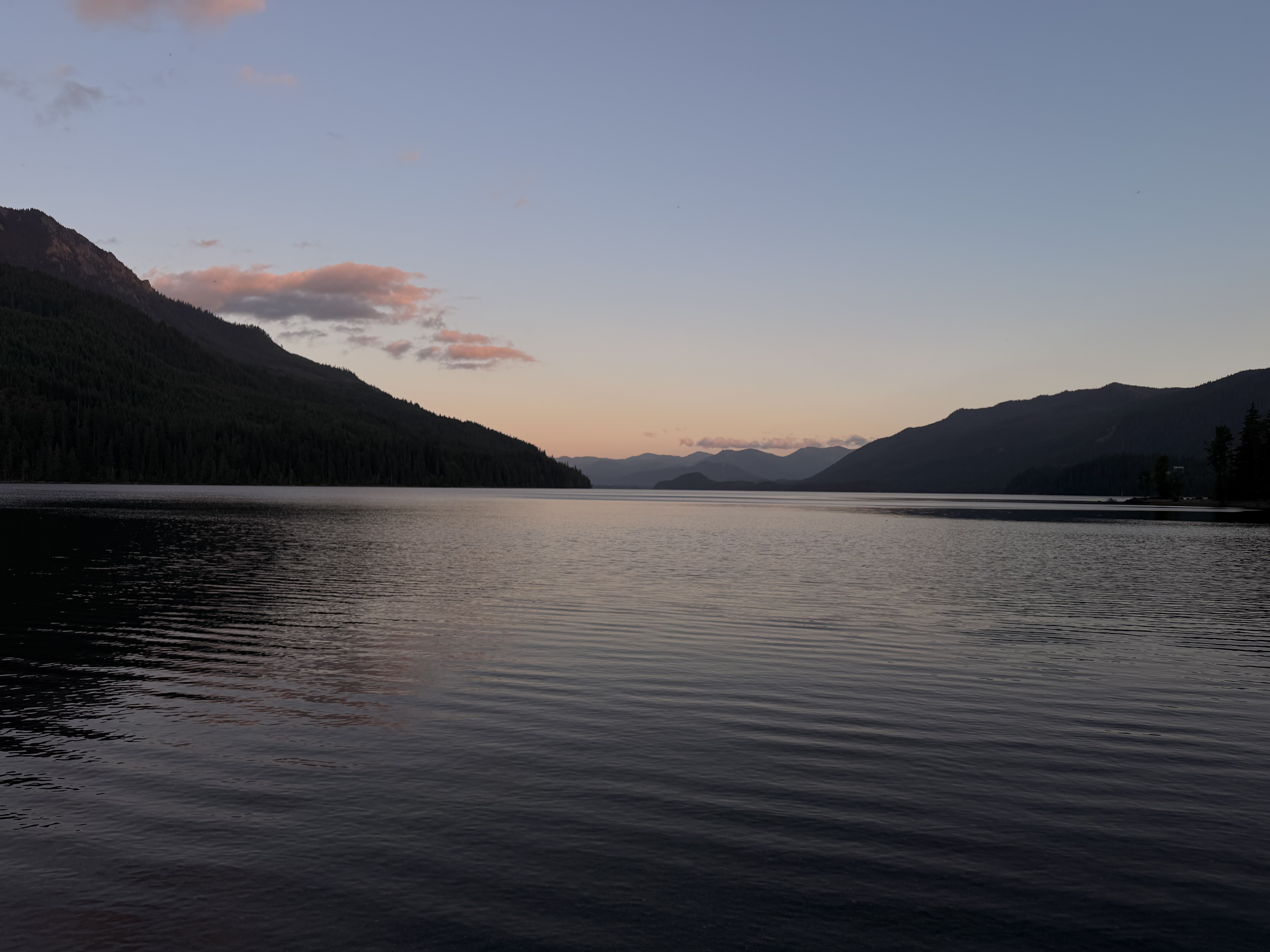 A tranquil alpine lake at twilight is framed by forested mountain ridges beneath a clear sky with soft pink clouds reflecting on the water's mirror-smooth surface.