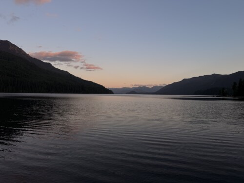 A tranquil alpine lake at twilight is framed by forested mountain ridges beneath a clear sky with soft pink clouds reflecting on the water's mirror-smooth surface.