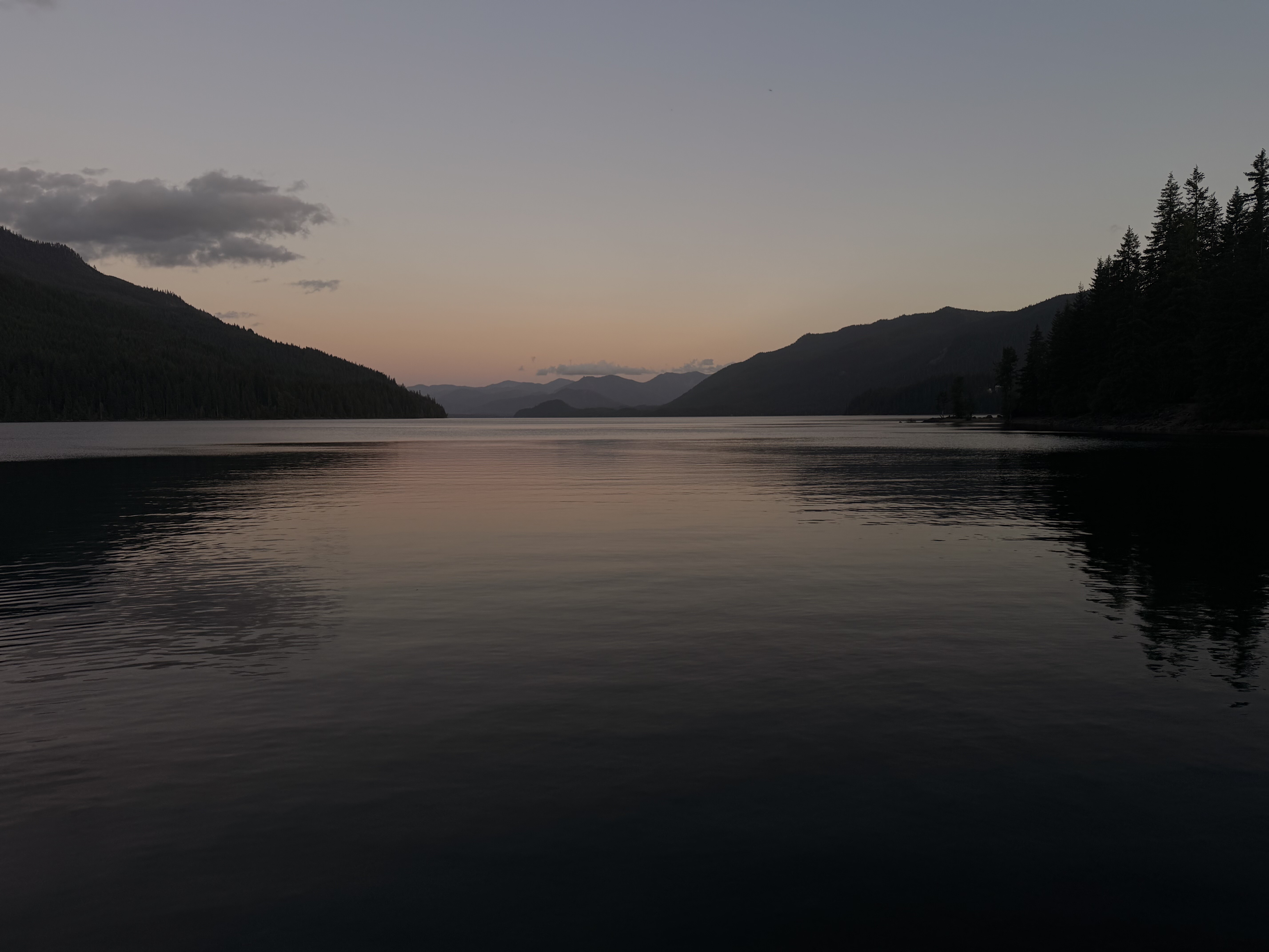 A calm alpine lake surrounded by forested mountains reflects a dusky sky with orange and purple tones near sunset.