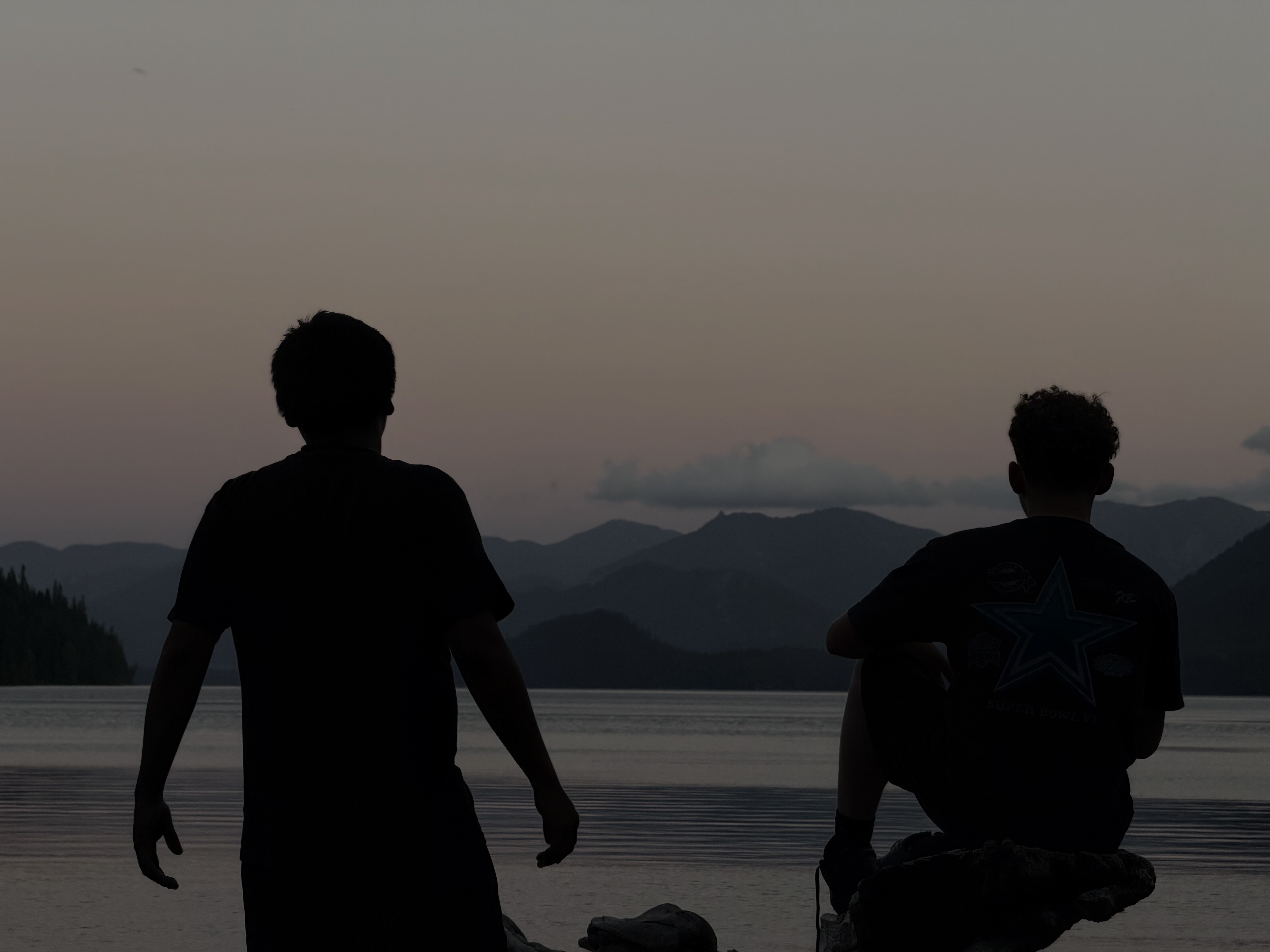 Two silhouetted figures stand in calm water facing a mountain-lined shoreline at dusk, with one walking and one crouching.