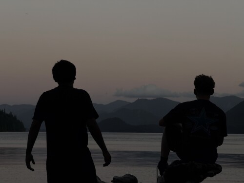 Two silhouetted figures stand in calm water facing a mountain-lined shoreline at dusk, with one walking and one crouching.