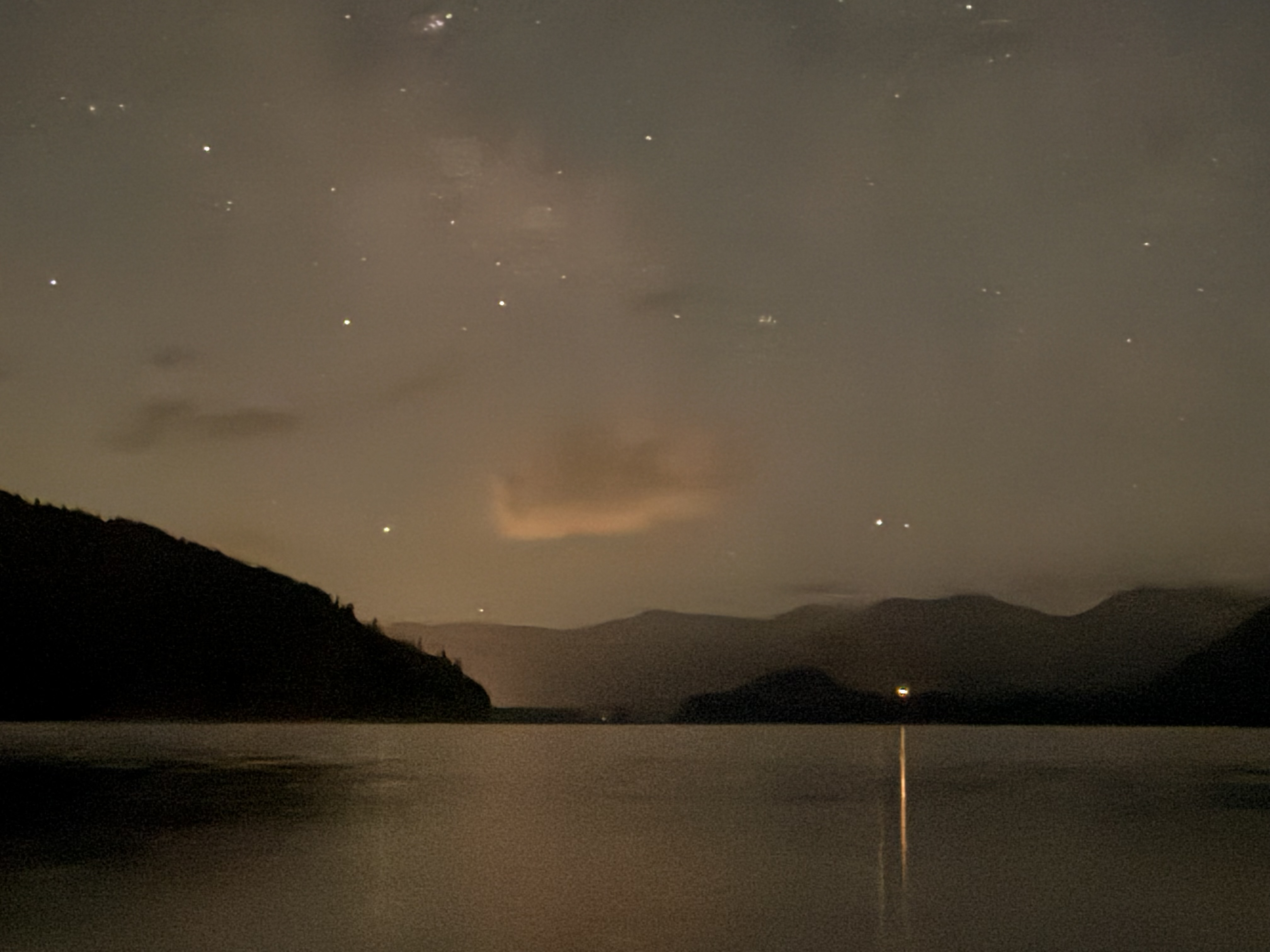 A vertical beam of light rises from a calm lake at twilight, with silhouetted mountains on the horizon and stars scattered across the darkening sky.