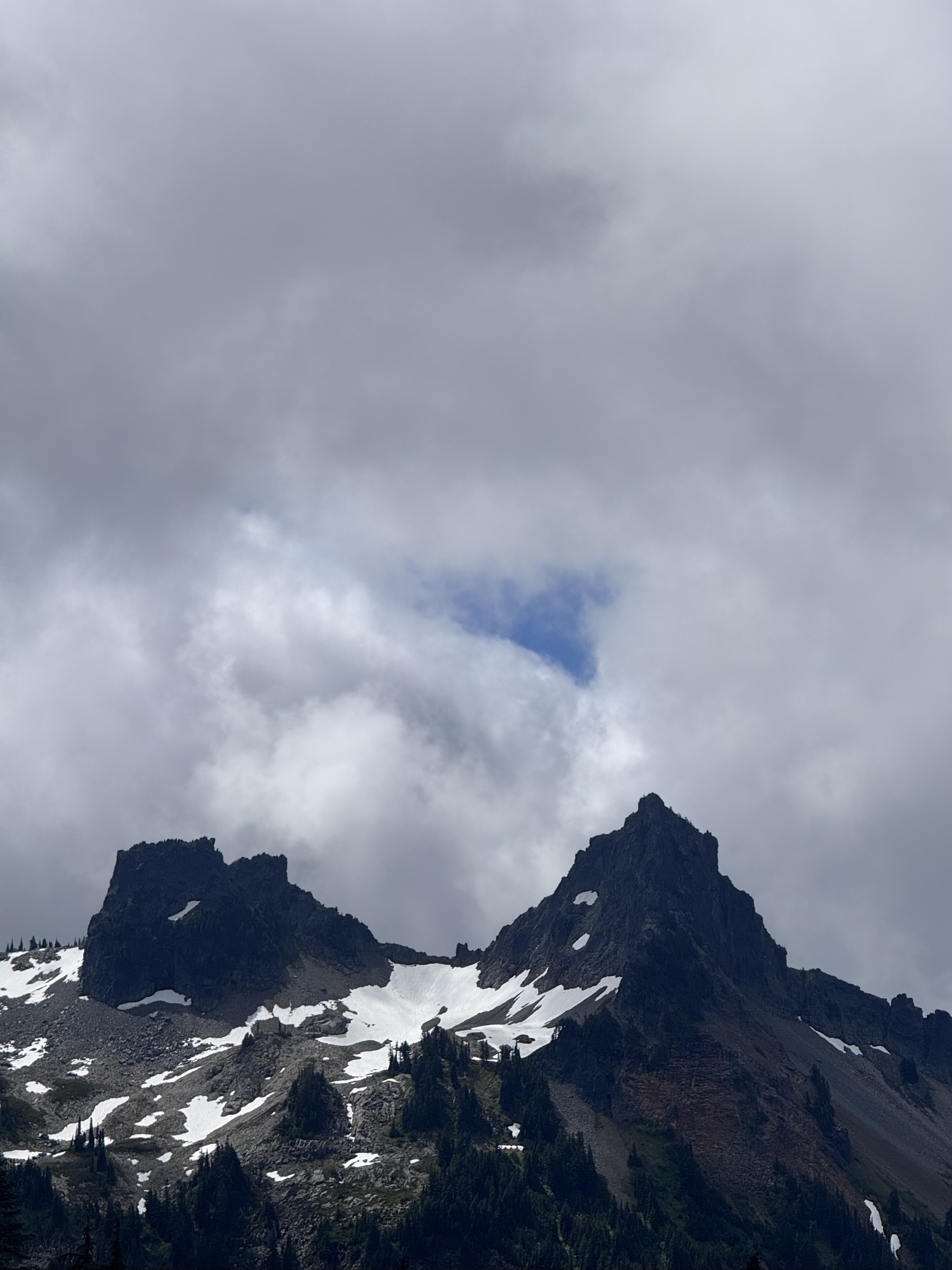 Two jagged mountain peaks with snow patches are silhouetted against a dramatic cloudy sky with a small break of blue in the center.