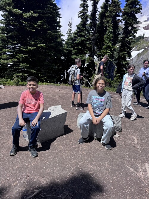 A group of children and adults gathered at a mountain viewpoint surrounded by tall evergreen trees, with two children seated in the foreground while others stand behind them.
