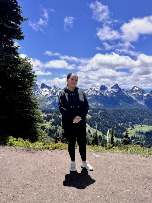A person in black athletic wear stands on a mountain overlook with snow-capped peaks, forested valleys, and blue sky with white clouds in the background.