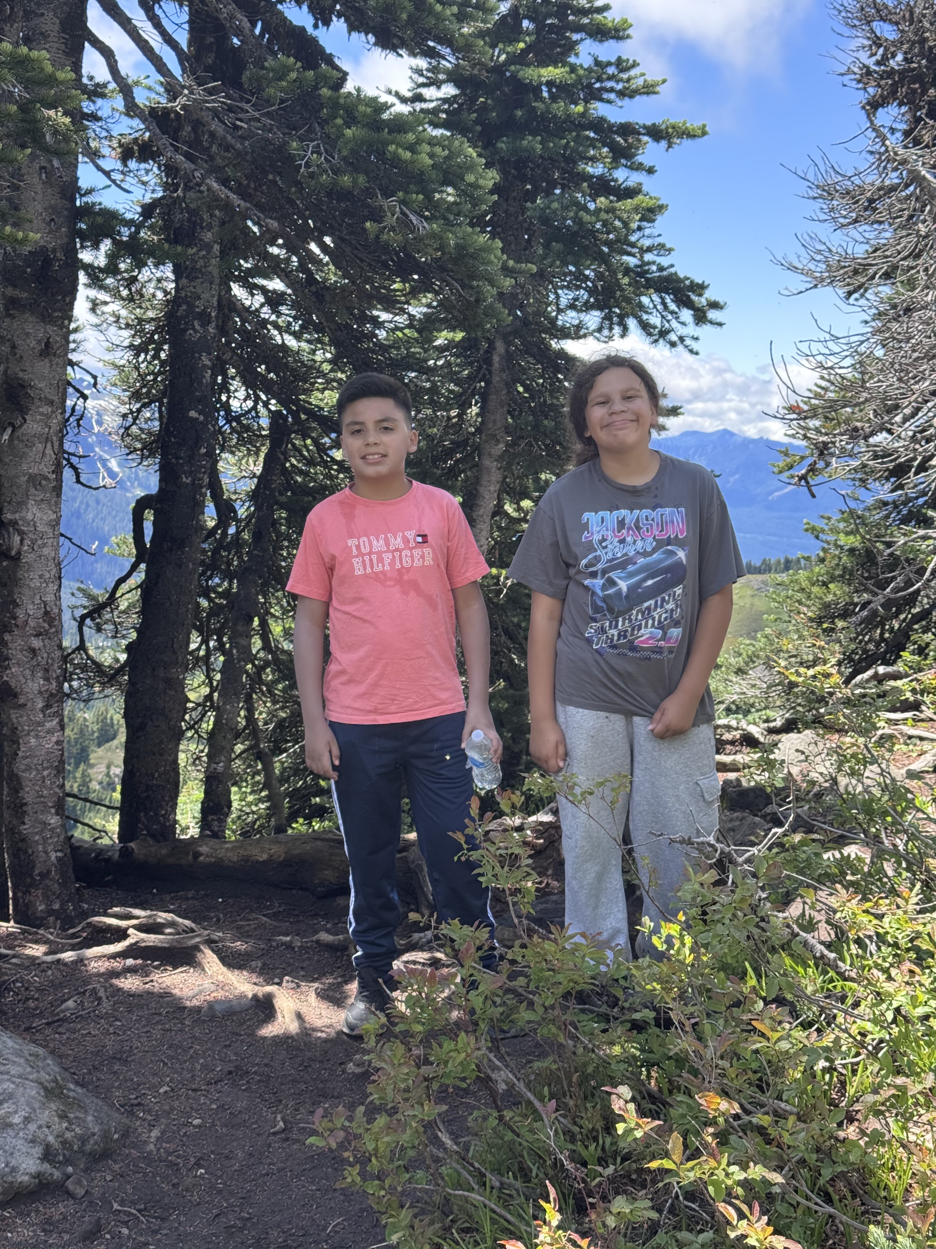 Two young people stand on a forest path between tall evergreen trees with mountains visible in the distance under a blue sky.