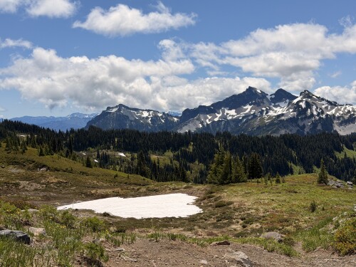 Alpine meadow with patches of lingering snow surrounded by coniferous forest, backed by jagged snow-capped mountain peaks under a partly cloudy sky.
