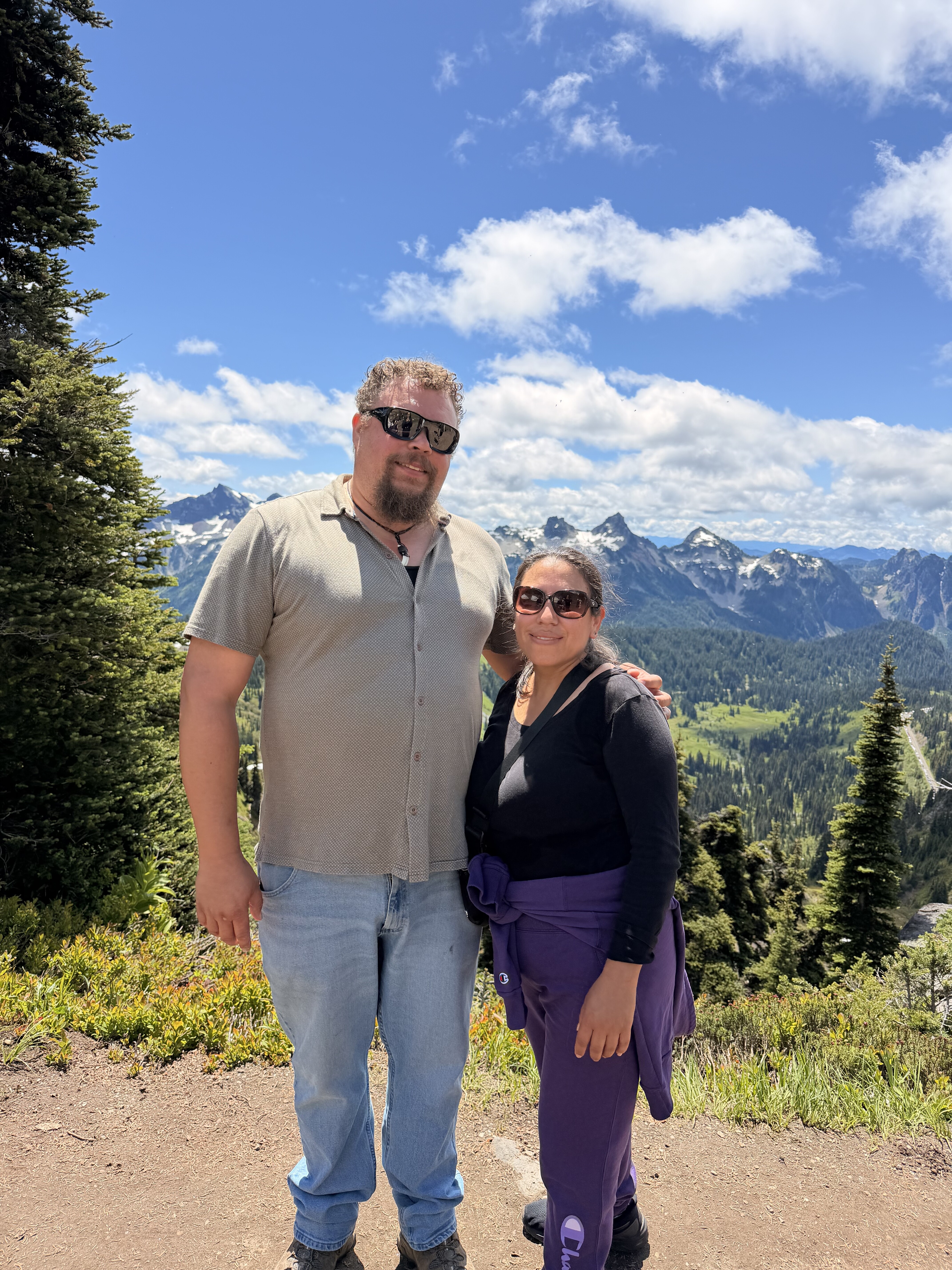 Two people stand together on a dirt mountain path with snow-capped peaks and forested slopes visible behind them under a partly cloudy sky.