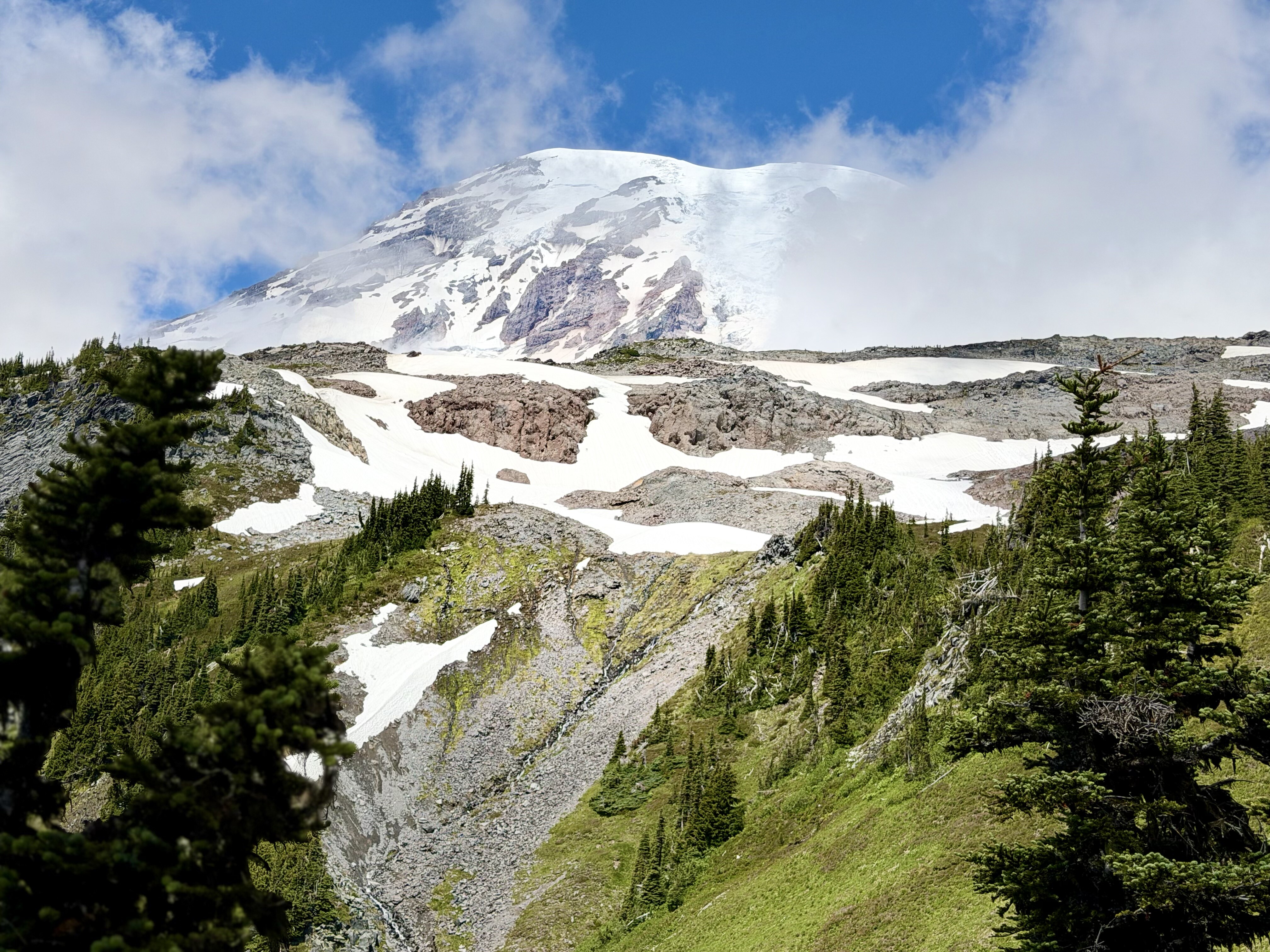 A snow-capped volcanic mountain rises above a forested valley dotted with patches of white snow and green evergreen trees under a partly cloudy blue sky.