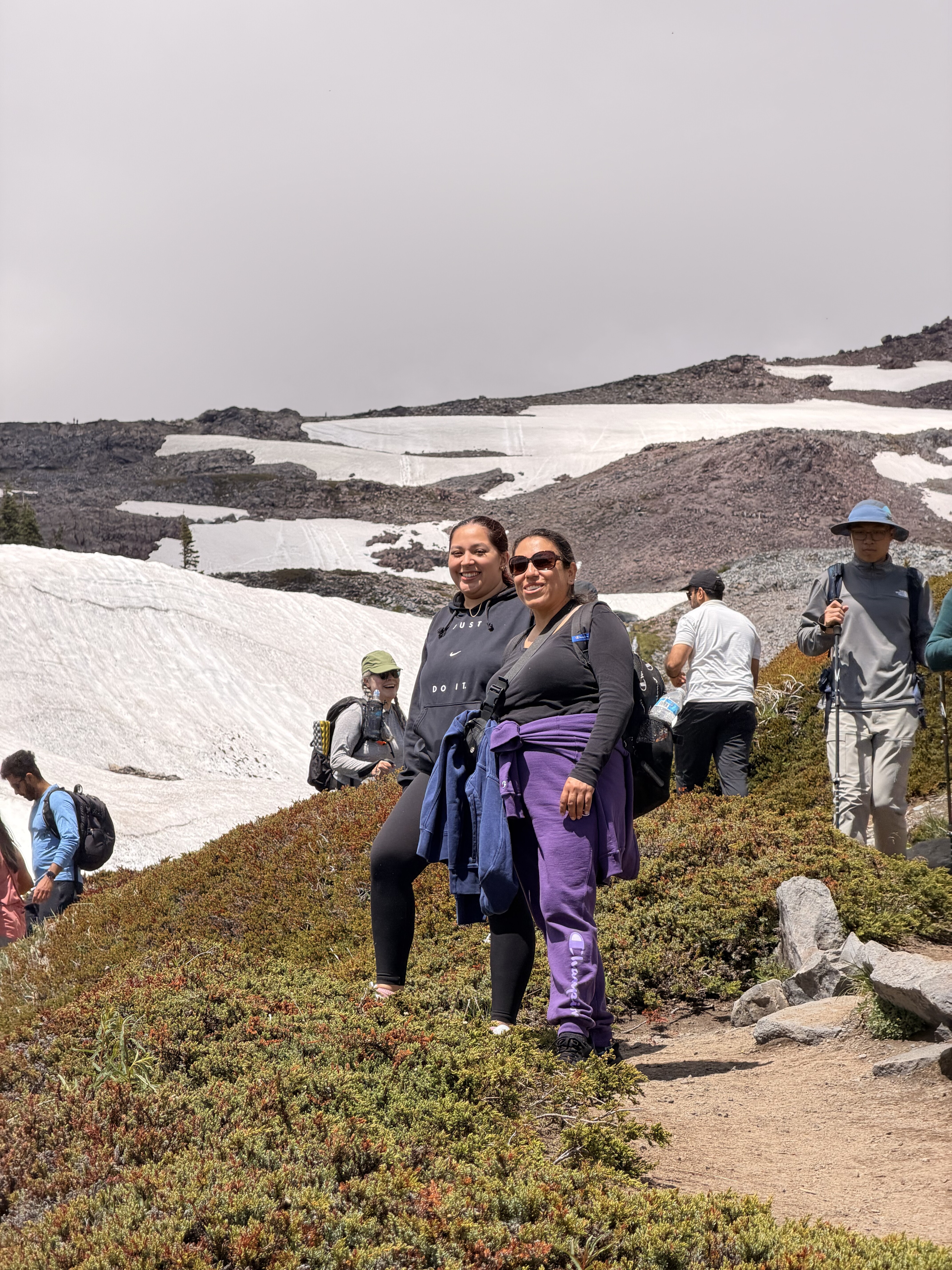 A group of hikers trek along a high-altitude mountain trail surrounded by patches of snow, rocky terrain, and low vegetation under an overcast sky.