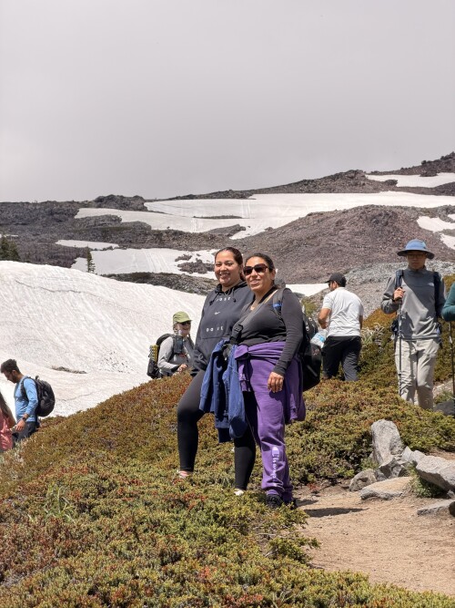 A group of hikers trek along a high-altitude mountain trail surrounded by patches of snow, rocky terrain, and low vegetation under an overcast sky.