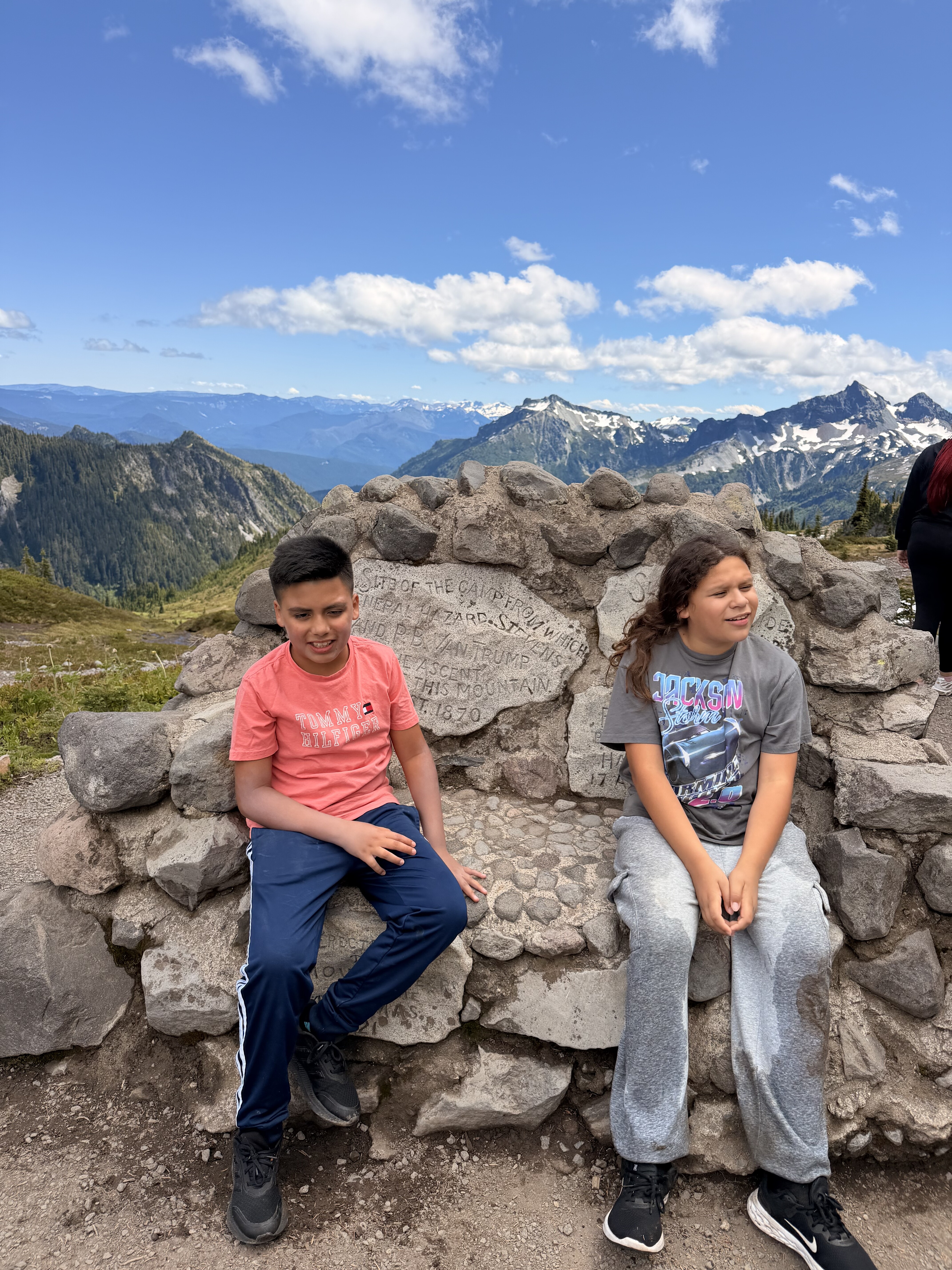A boy and girl sit on a large stone formation overlooking a scenic mountain landscape with snow-capped peaks, forested valleys, and a blue sky with white clouds.