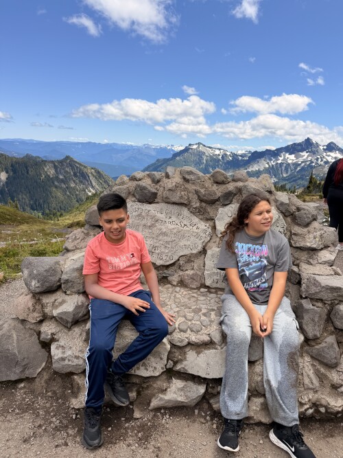 A boy and girl sit on a large stone formation overlooking a scenic mountain landscape with snow-capped peaks, forested valleys, and a blue sky with white clouds.
